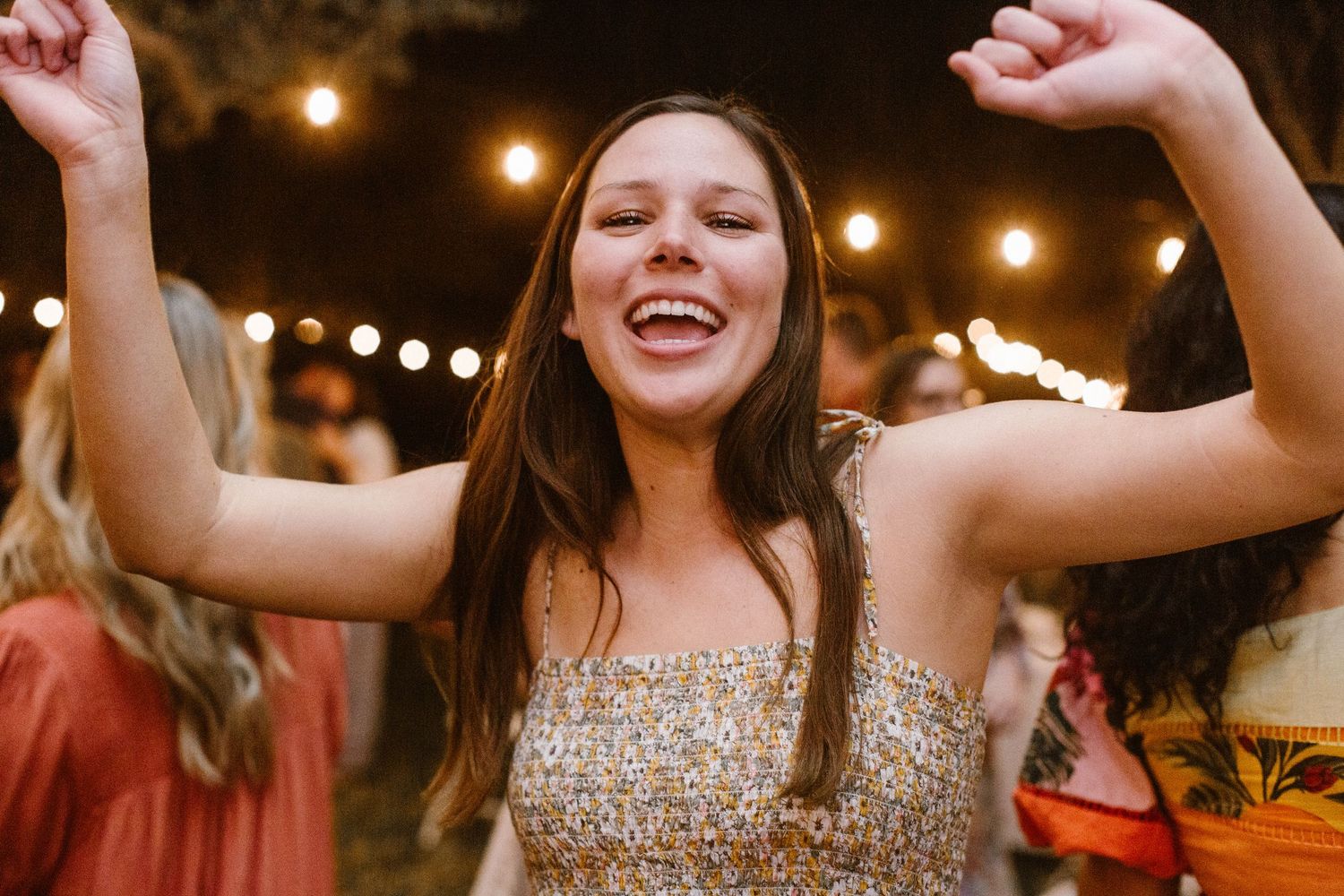 Young woman in floral dress dancing joyfully under string lights at an outdoor party.
