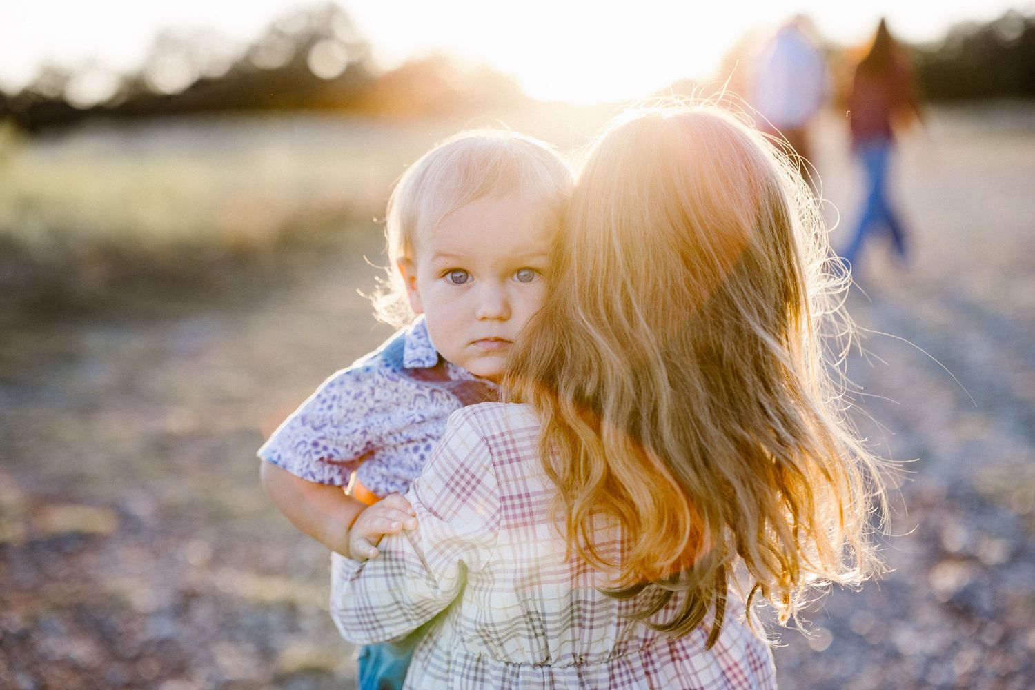 A woman with curly hair holds a baby outdoors in warm golden sunset light.