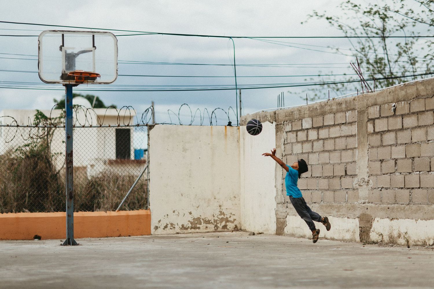 A young boy leaps to shoot a basketball on an outdoor court with a concrete wall backdrop.