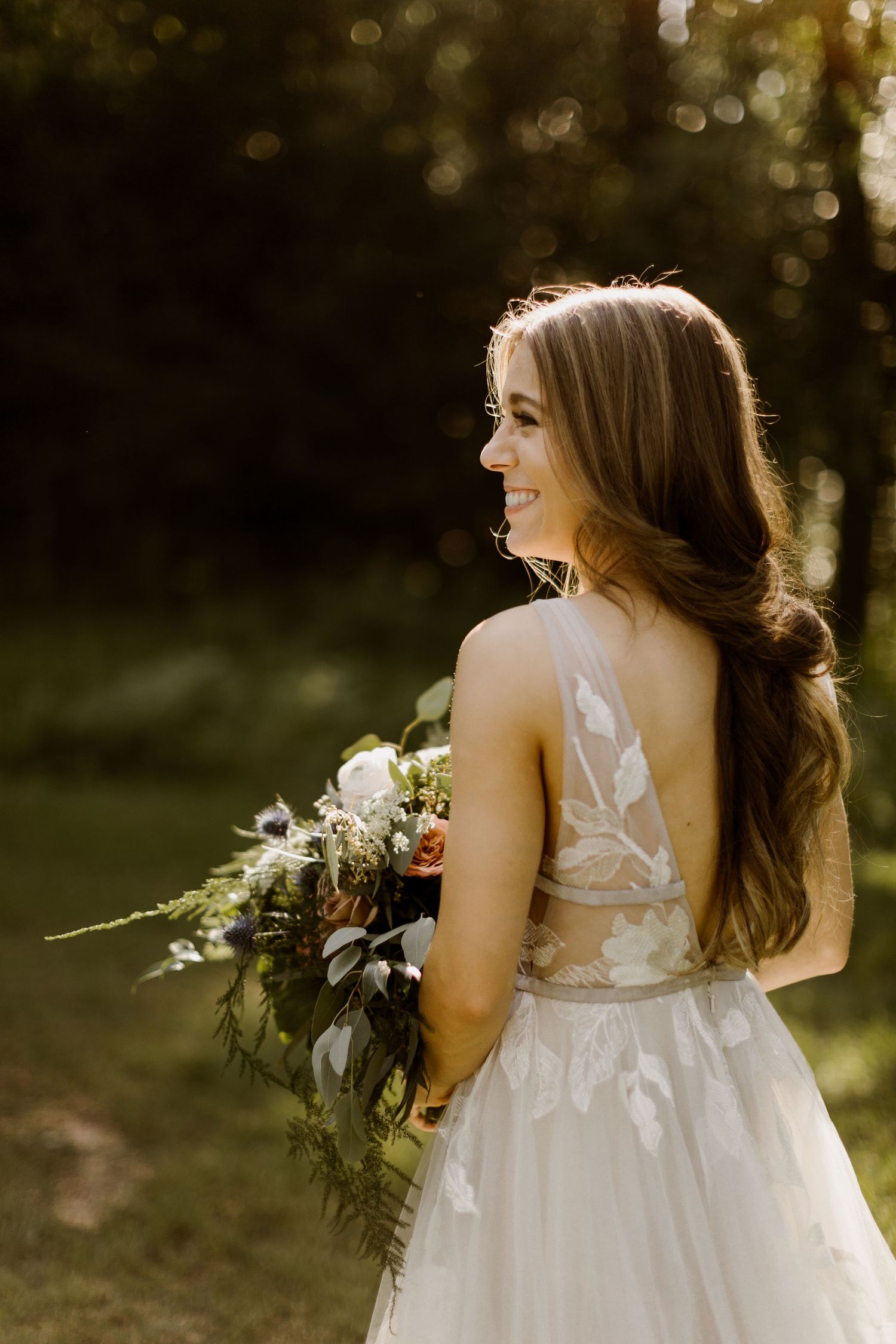 Bride in lace backless wedding gown holds wildflower bouquet while smiling in golden hour forest light.