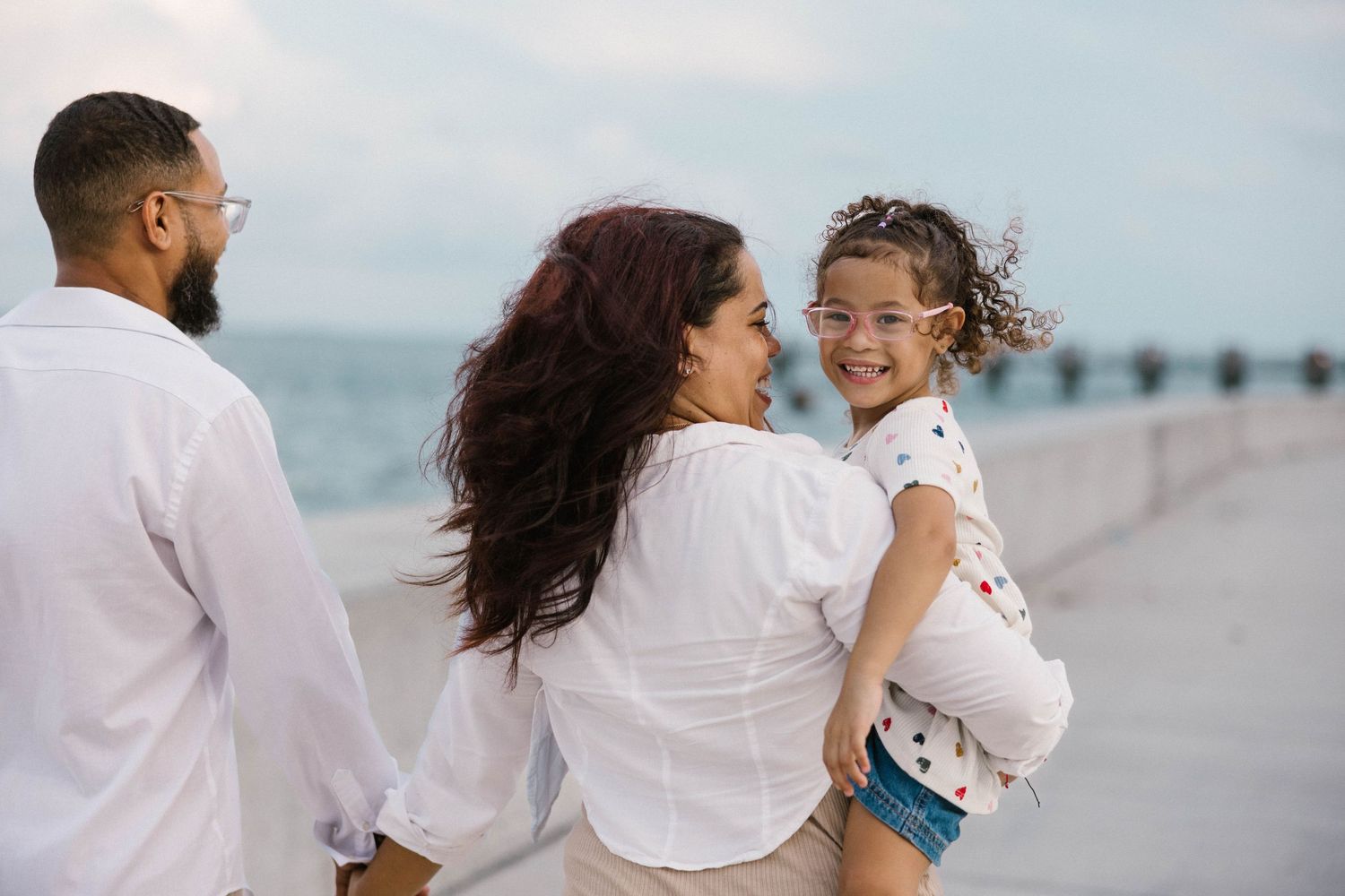 A woman holds her young daughter while walking with a man along a seaside promenade at dusk.
