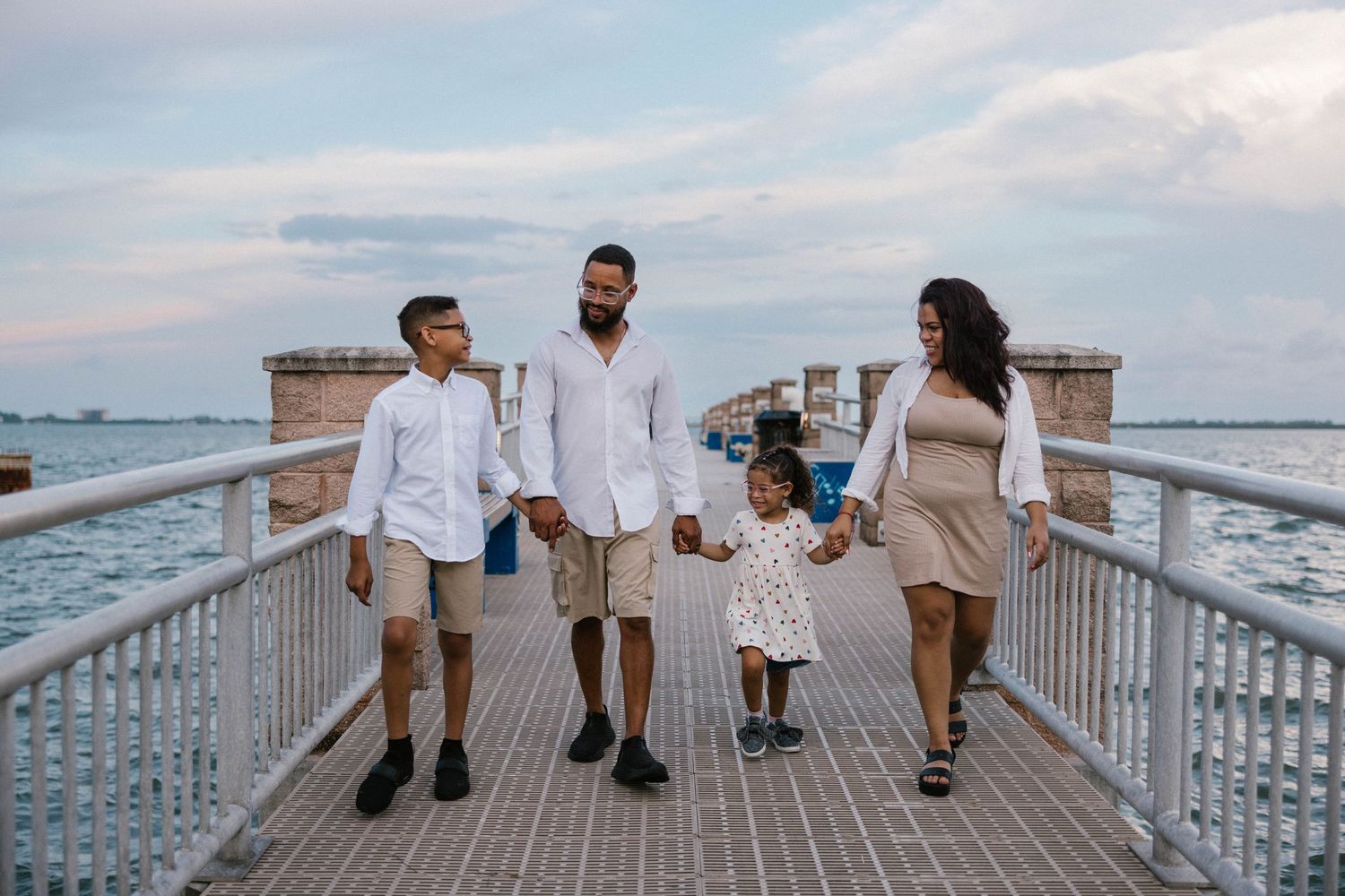Family of four holding hands walking on a waterfront pier at sunset in coordinating white and neutral outfits.