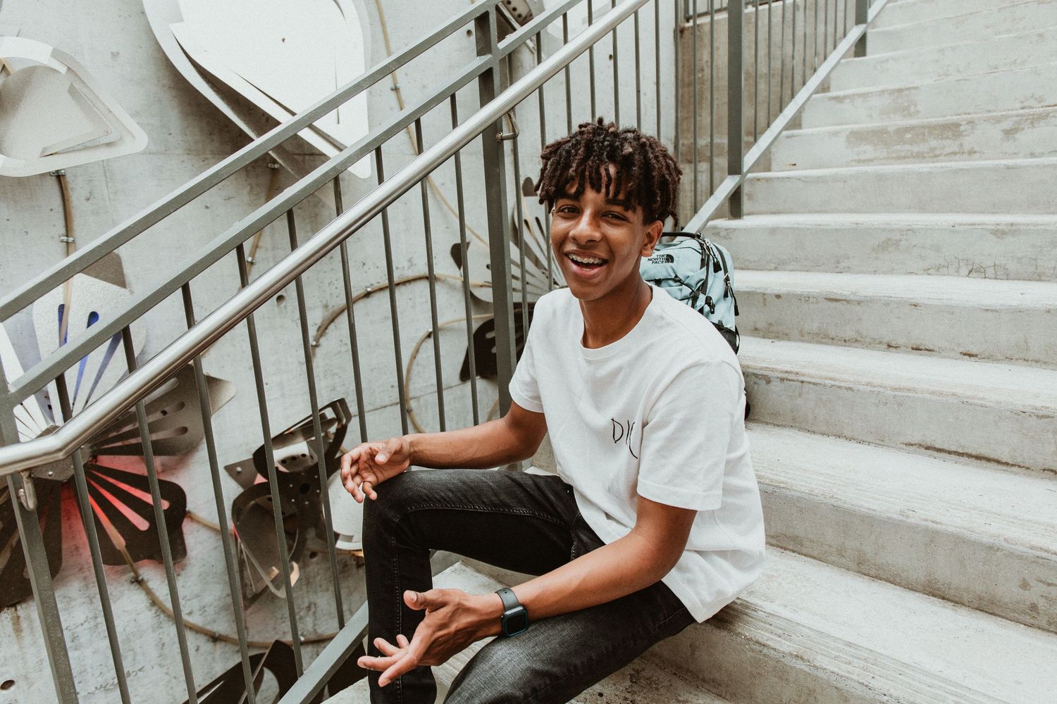 Young man with curly hair smiling while sitting on outdoor stairs wearing a white t-shirt and dark jeans.
