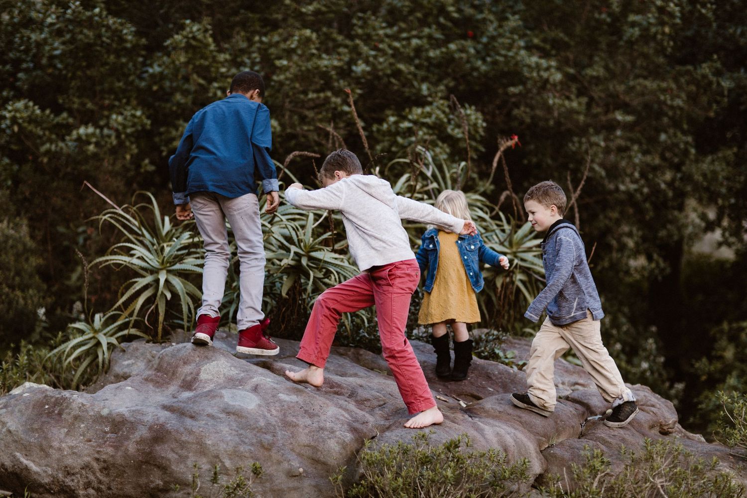 Four children helping each other climb a large rock surrounded by lush green foliage outdoors.