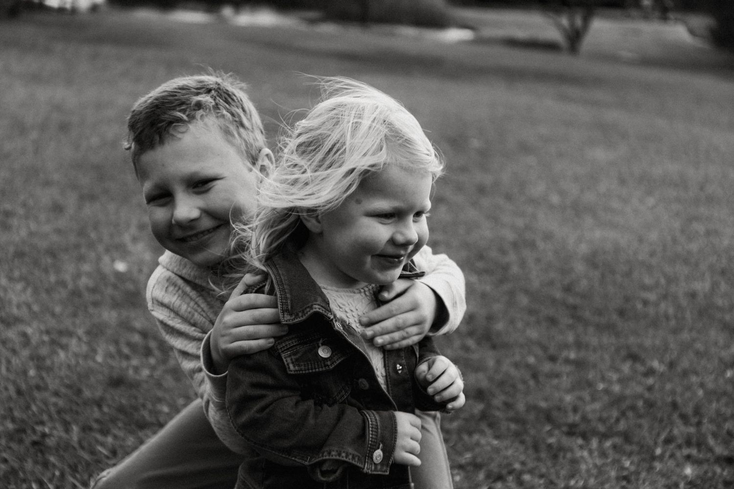 Black and white photo of two young siblings hugging and laughing together outdoors on green grass.