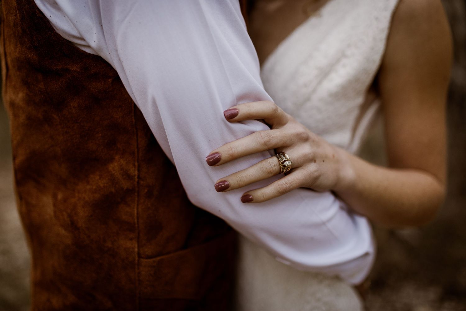 Close-up of bride's hand with gold ring resting on groom's arm in white shirt and brown suede vest.
