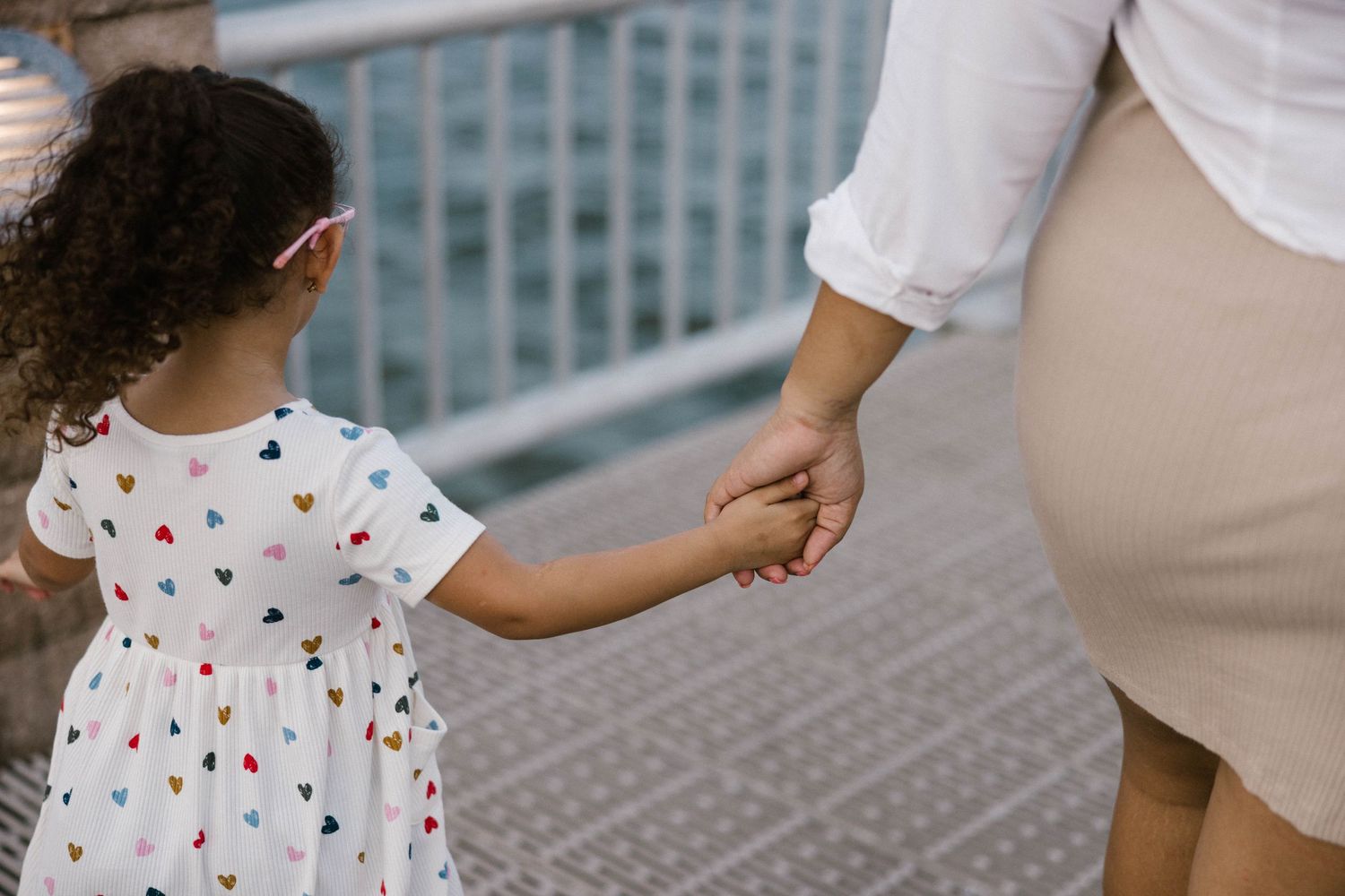 Young girl in a heart-print dress holding hands with her mother while walking along an outdoor walkway.