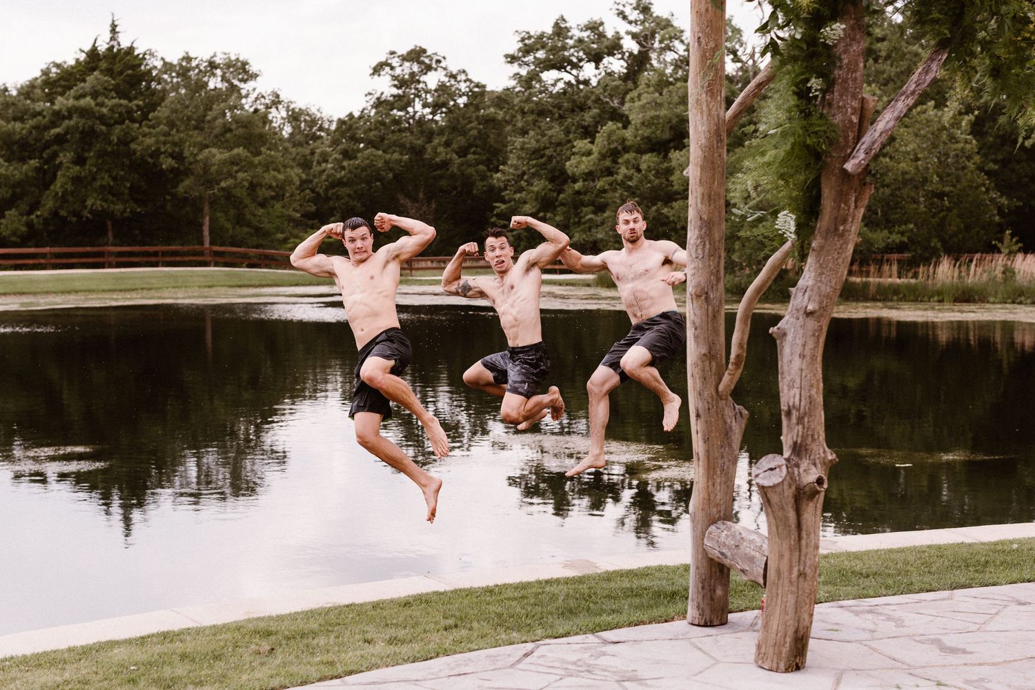 Four groomsmen jumping joyfully into lake surrounded by lush green trees.