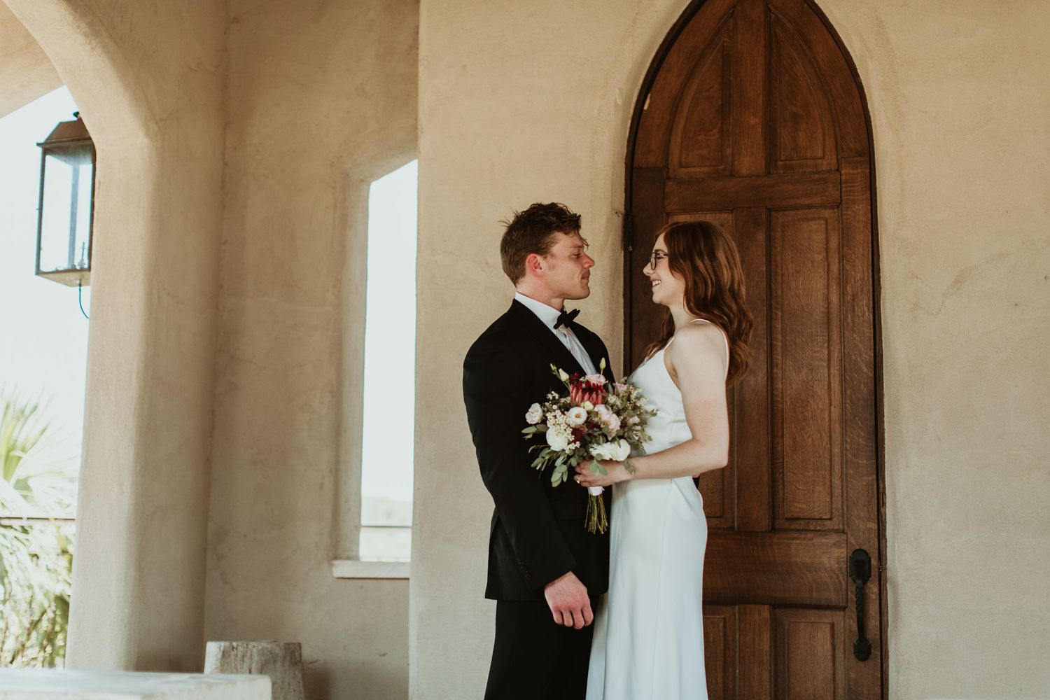 Bride and groom stand together holding a floral bouquet before an arched wooden door at an elegant wedding venue.