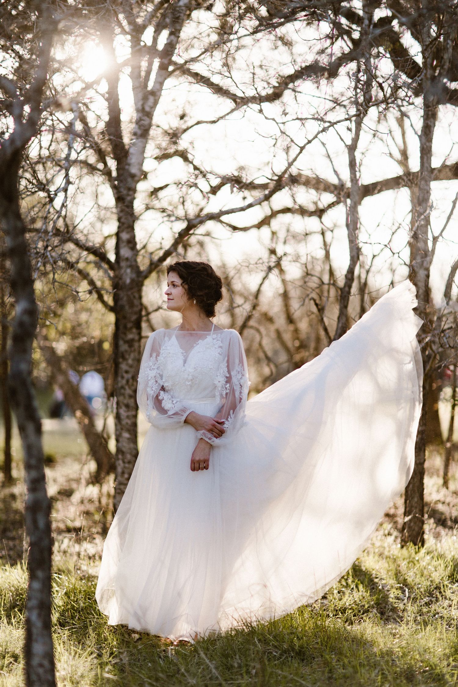 Bride in flowing white lace wedding dress spinning among bare trees in golden sunset light.