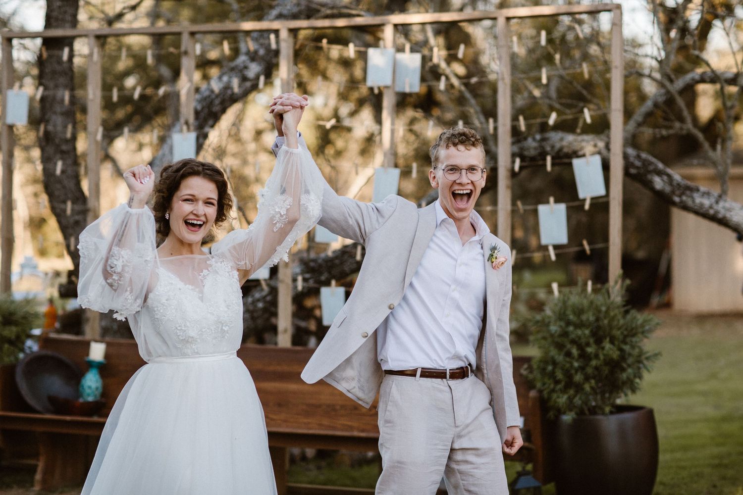 Joyful bride and groom raising hands in celebration after outdoor wedding ceremony under string lights.