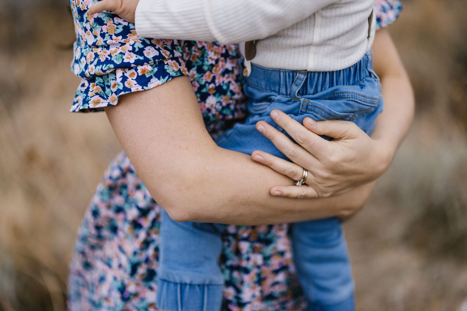 Close-up of a mother holding a child in blue jeans, wearing a floral dress and engagement ring.