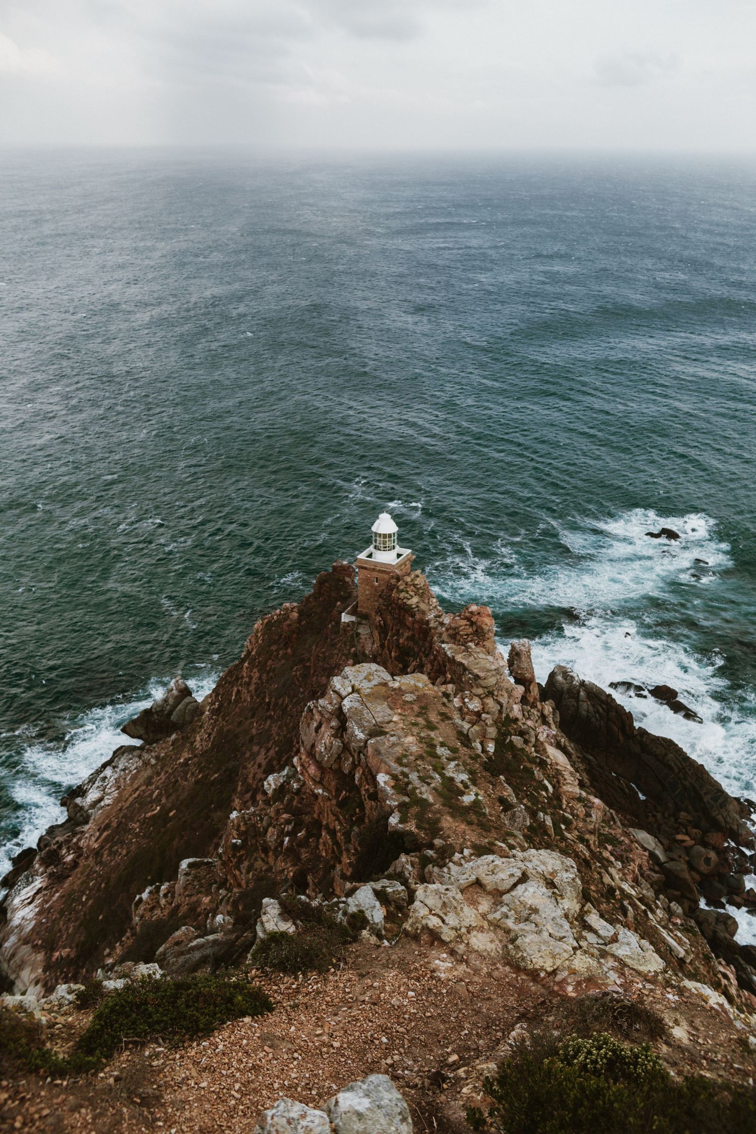 Aerial view of a small white lighthouse perched on a rocky coastal cliff above a vast green ocean.