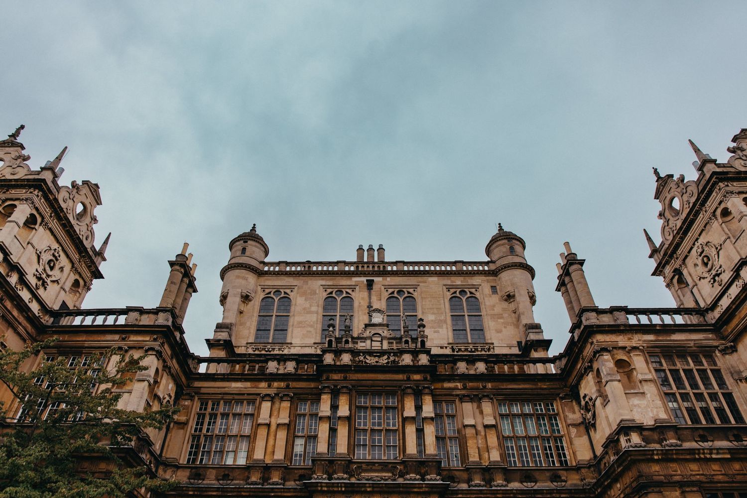 Low-angle view of ornate Gothic architecture with turrets and large windows under cloudy sky.