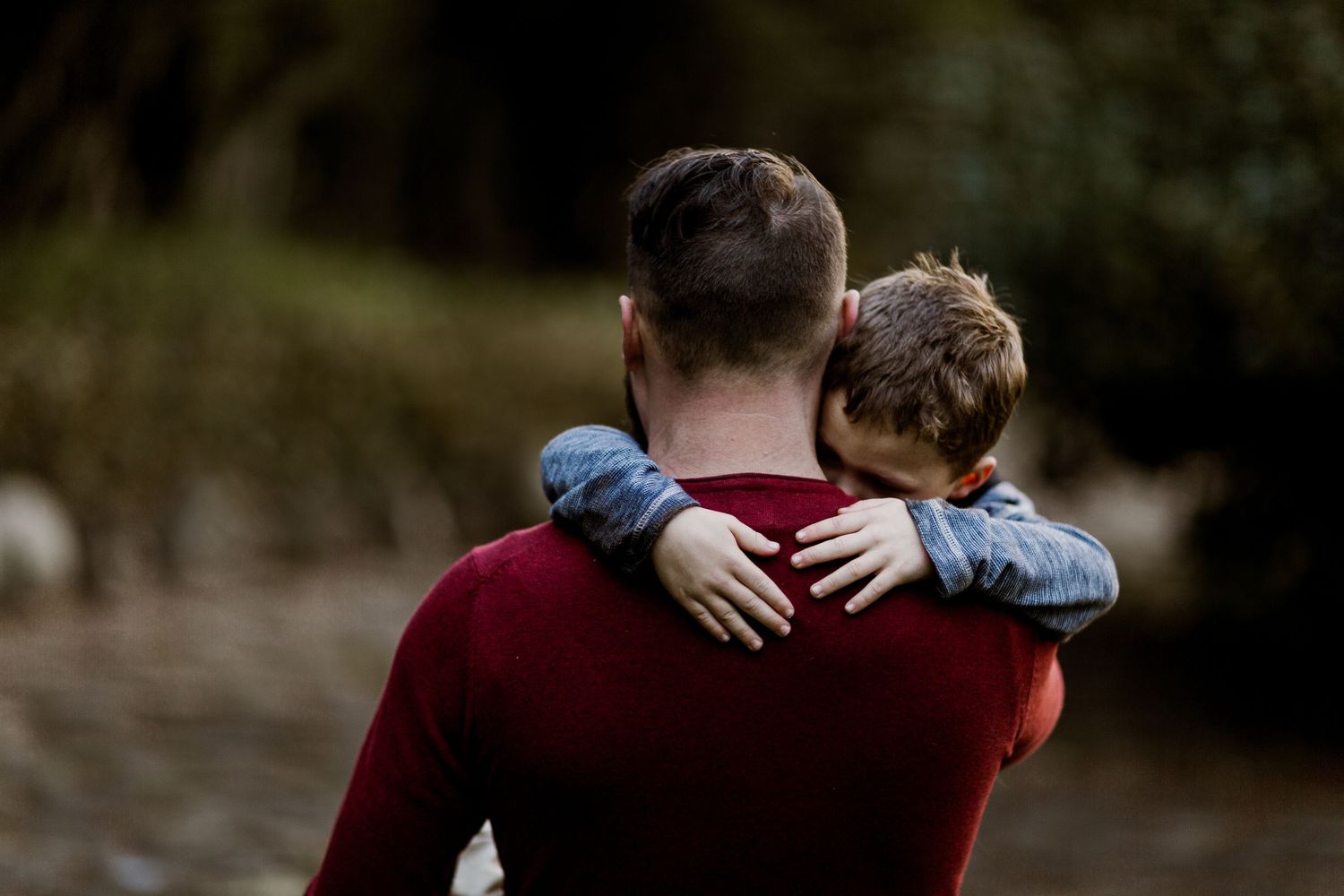 Father carrying young son on his back in a tender outdoor embrace from behind.