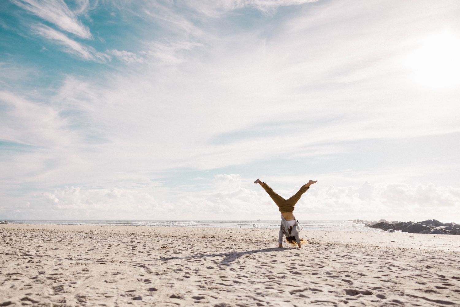 Person doing a handstand on a wide sandy beach under a blue sky with clouds.