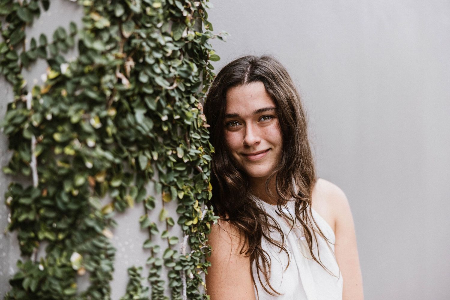 Brunette bride in white dress posing beside ivy-covered wall outdoors.