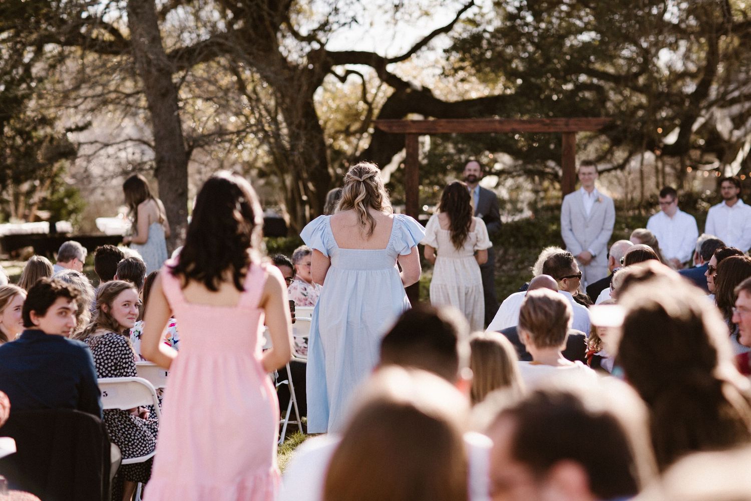 Wedding guests seated in white chairs watch bridesmaids in pastel dresses walk down an outdoor aisle toward a wooden arch.