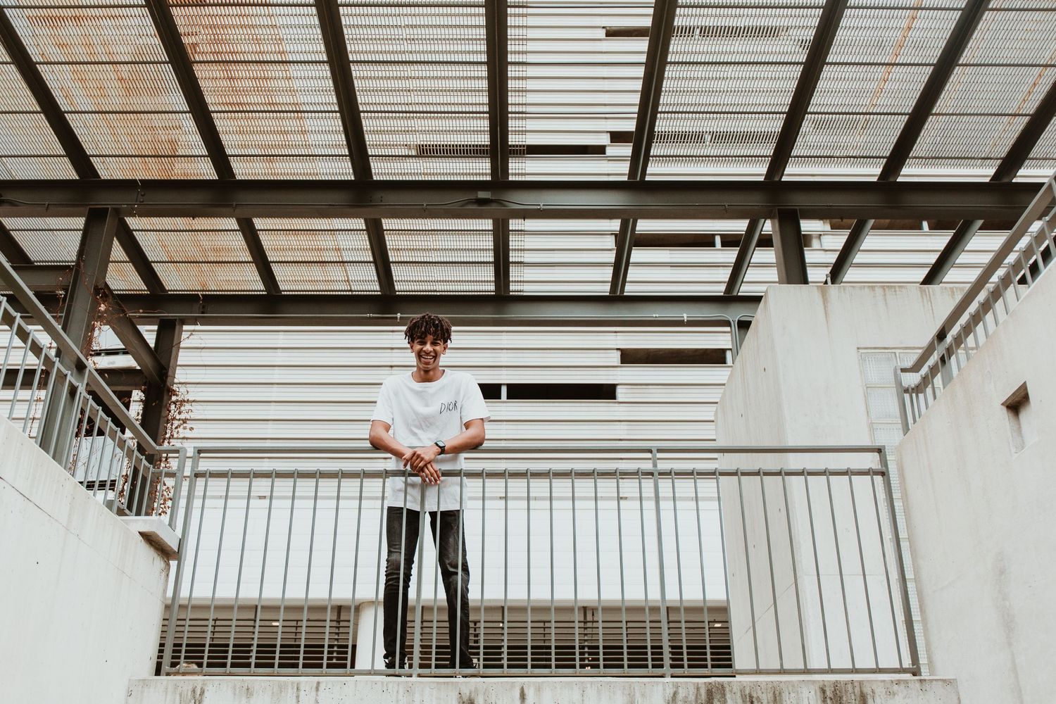 Young man in white t-shirt and black pants leaning on metal railing in modern industrial building.