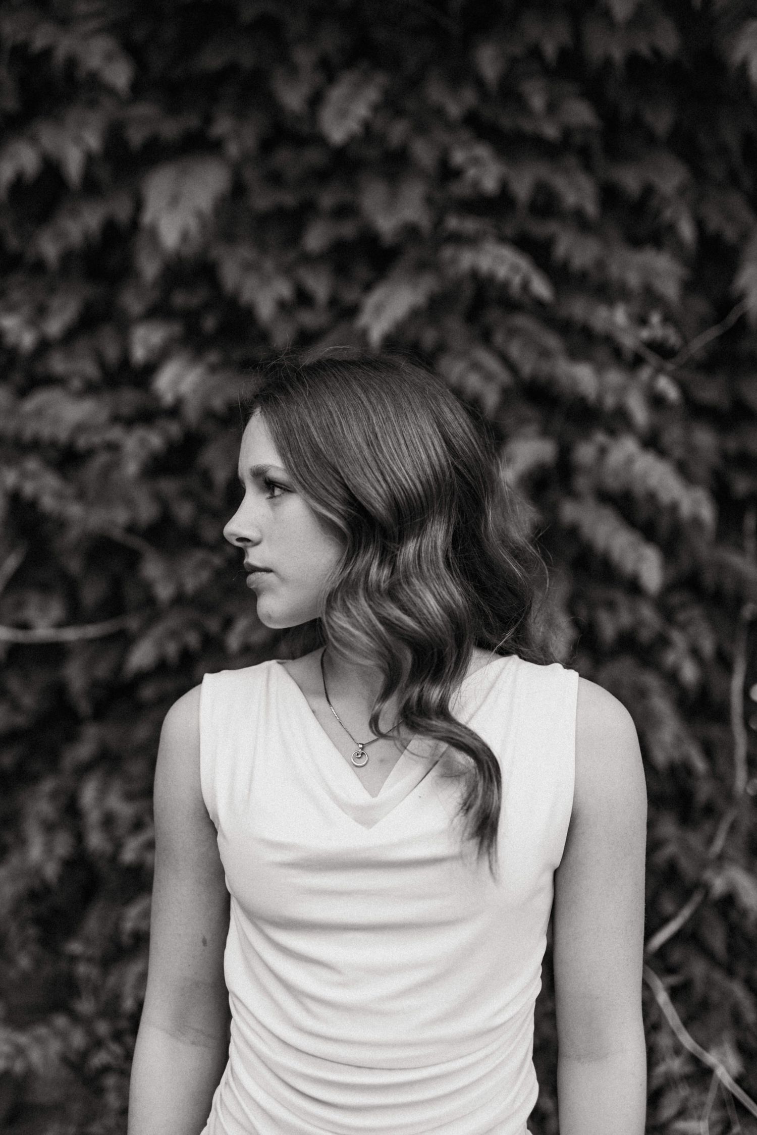 Black and white portrait of a woman in a white dress with wavy hair standing before a leafy backdrop.