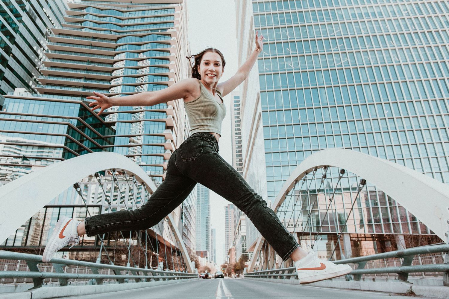 Athletic young woman jumping joyfully on urban bridge surrounded by modern glass skyscrapers.