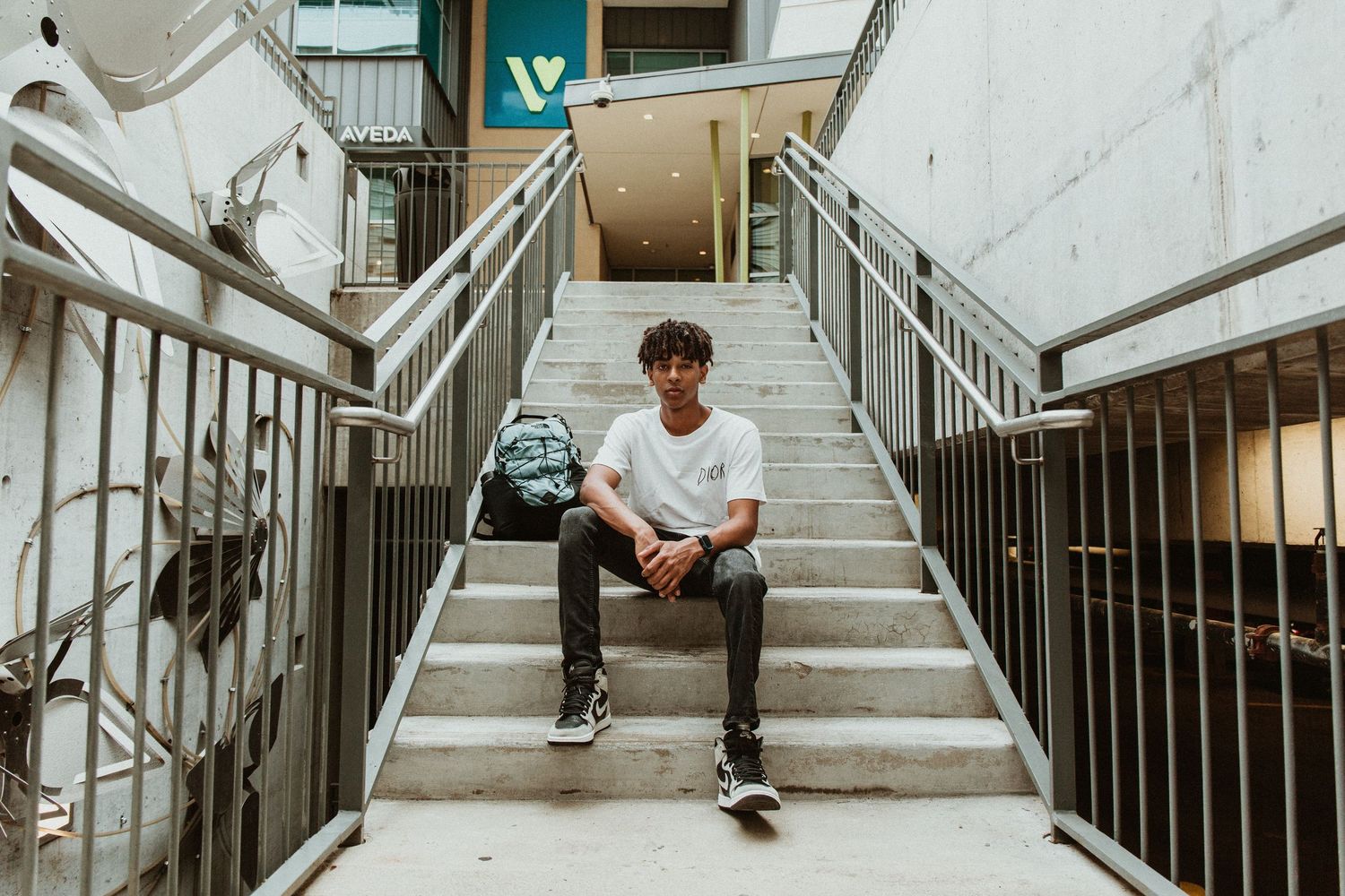 Young man sitting on concrete stairs with backpack in urban building setting.
