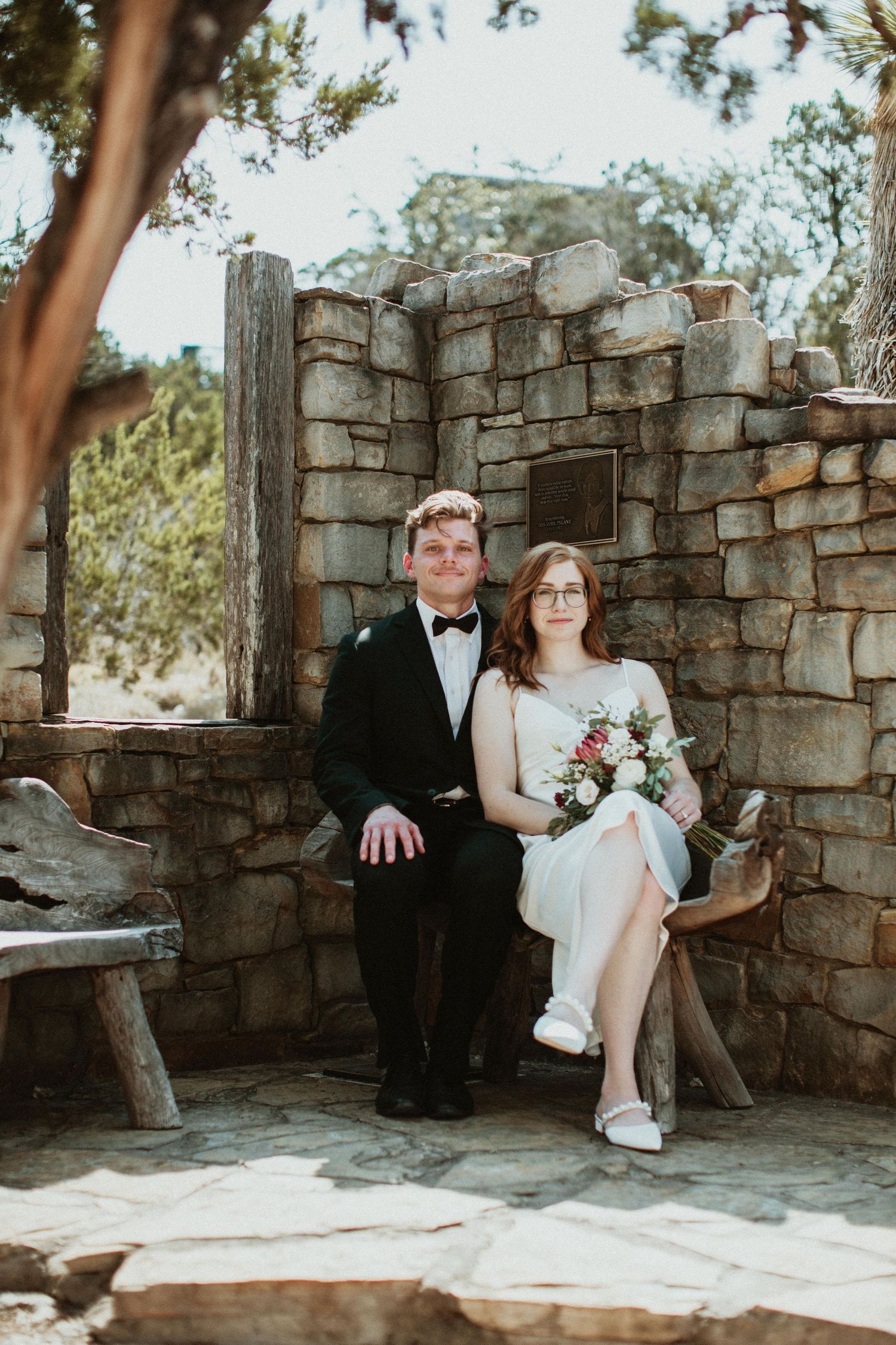 A bride and groom sit together in front of a stone wall outdoors, she holds a floral bouquet, both smiling at the camera.