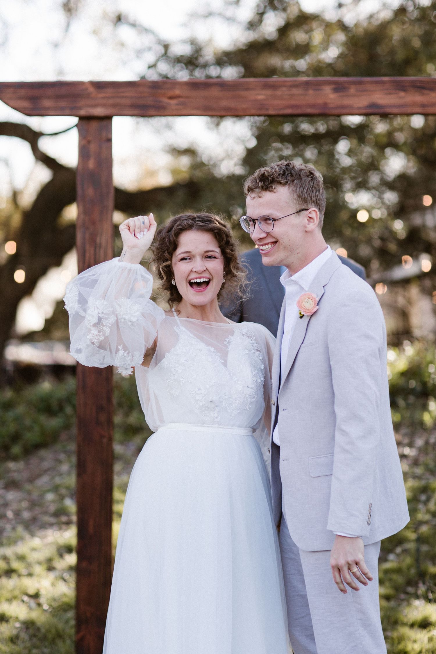 Excited bride pumps fist while laughing with groom in gray suit after outdoor wedding ceremony.