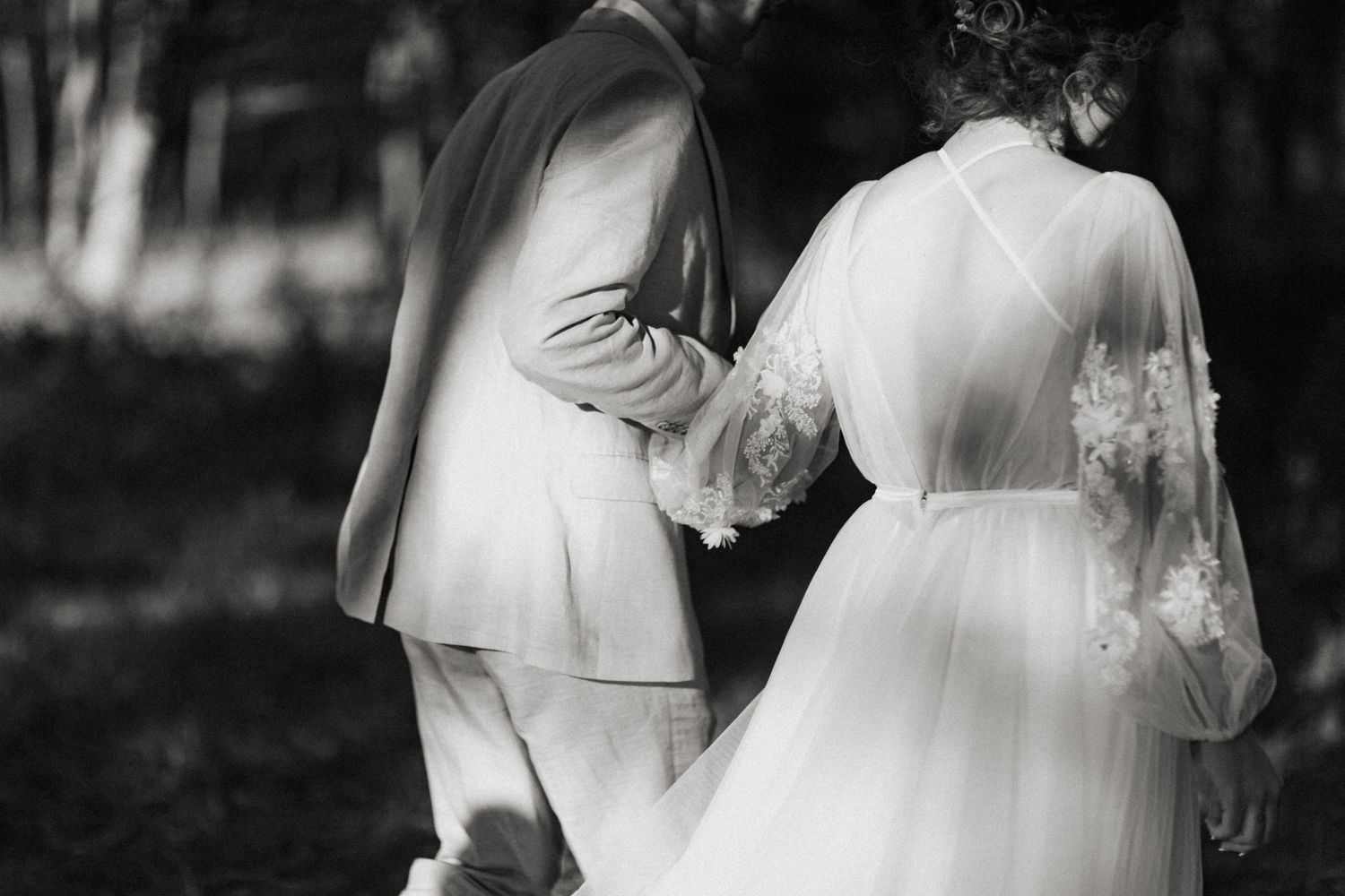 Black and white photo of bride in lace dress and groom in suit walking hand in hand outdoors.