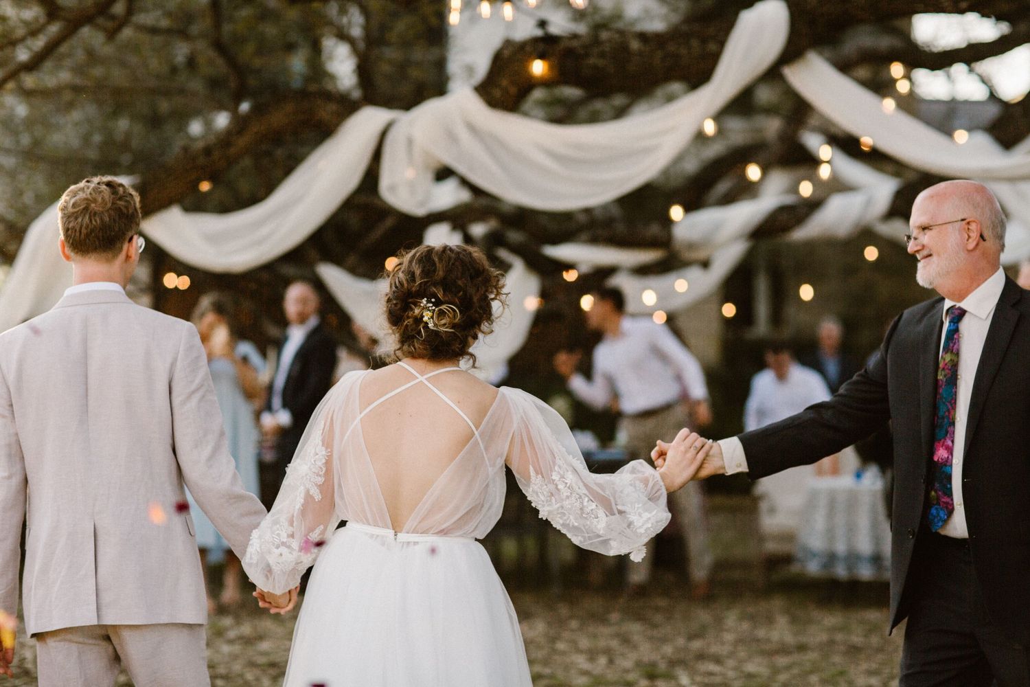 Bride dancing with older gentleman while groom looks on at outdoor wedding under white draped decorations.