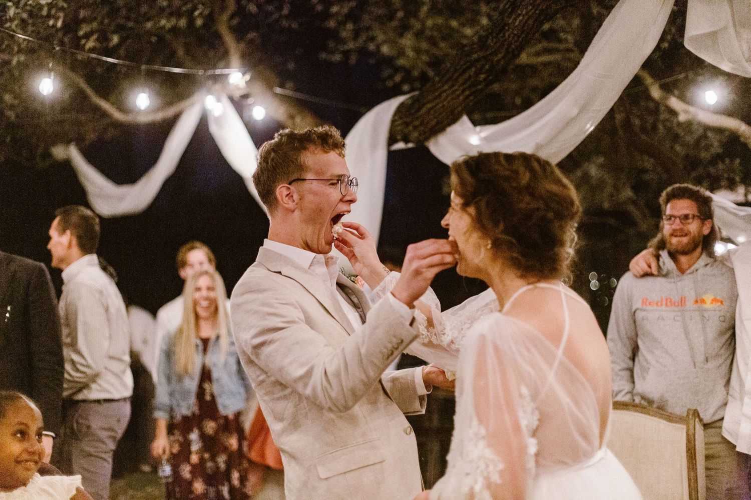 Bride and groom sharing wedding cake at outdoor evening reception under string lights.