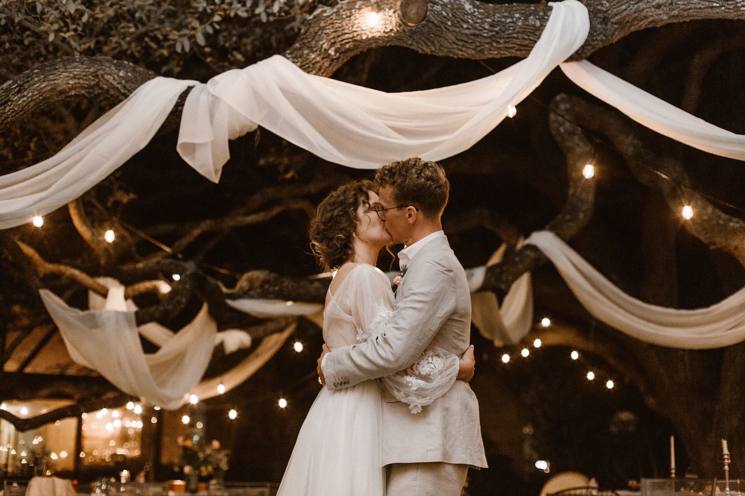 Bride and groom sharing romantic kiss under white drapes and string lights at night.