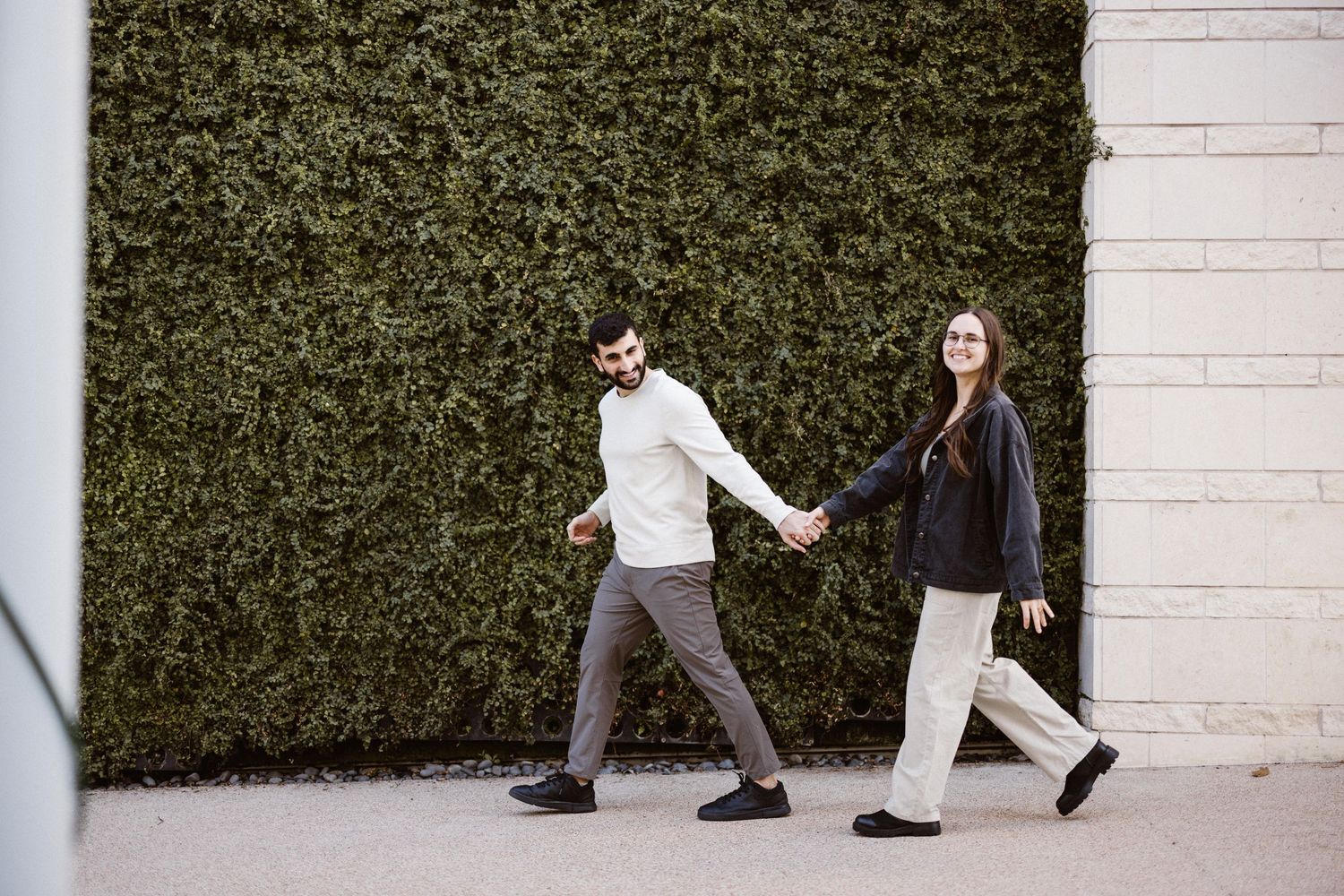 Happy couple holding hands walking in front of a lush green ivy wall outdoors.