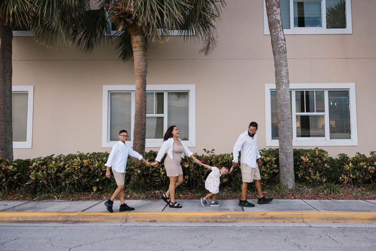 Family of four in white outfits holding hands and walking along a sunny urban sidewalk.