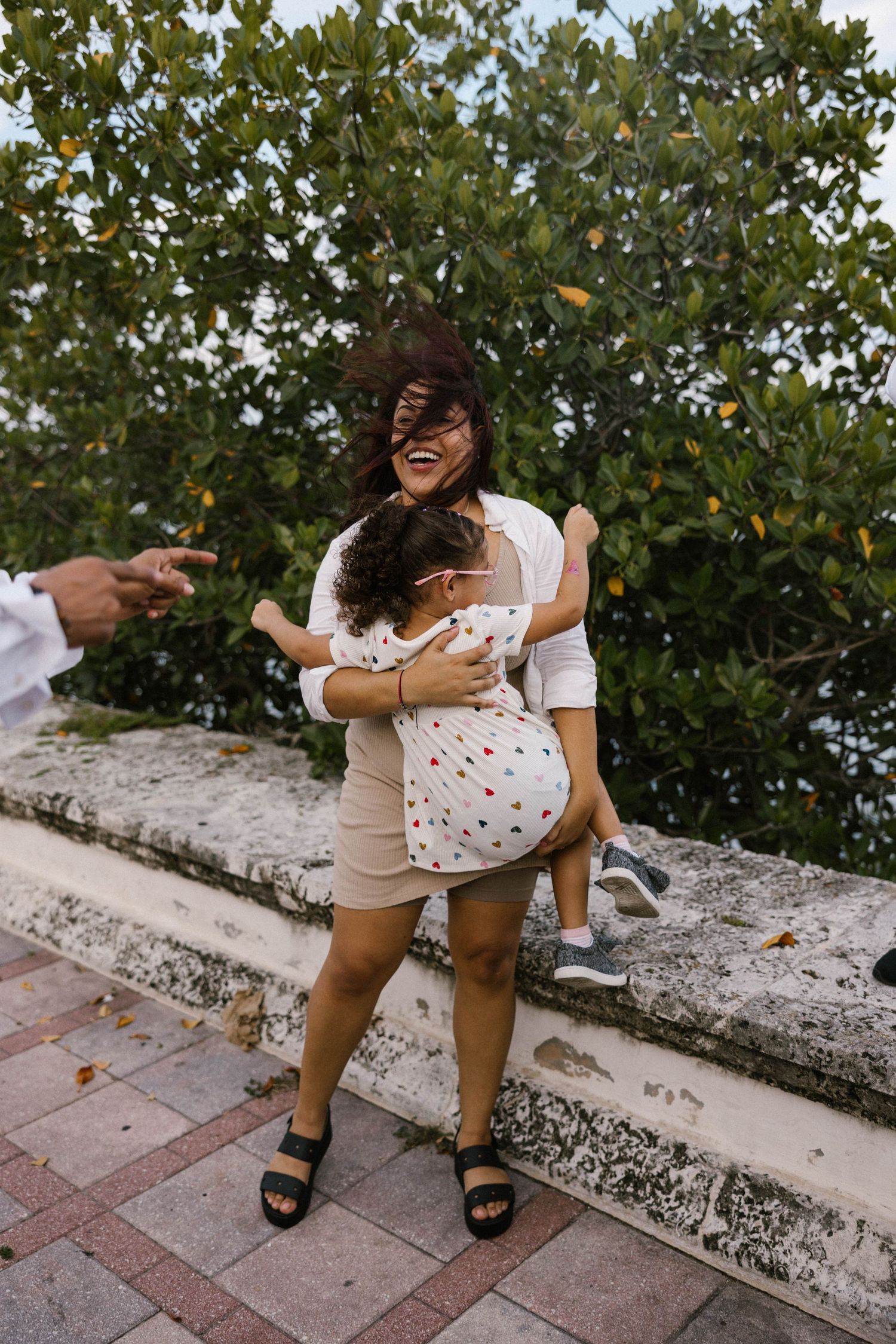 Laughing mother with windswept hair hugs her young child outdoors near green trees.