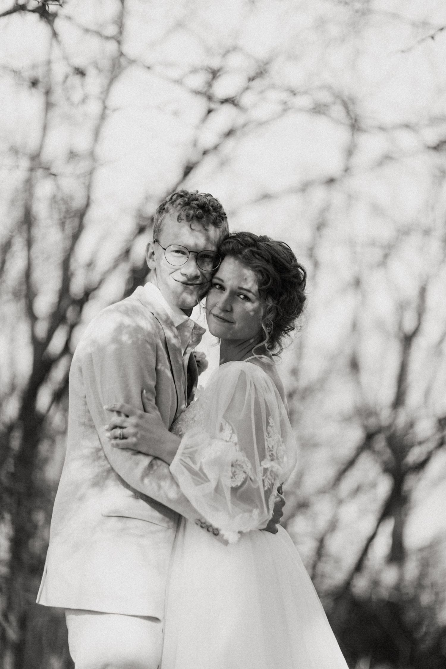 Black and white portrait of bride in lace gown and groom in suit embracing before bare winter trees.