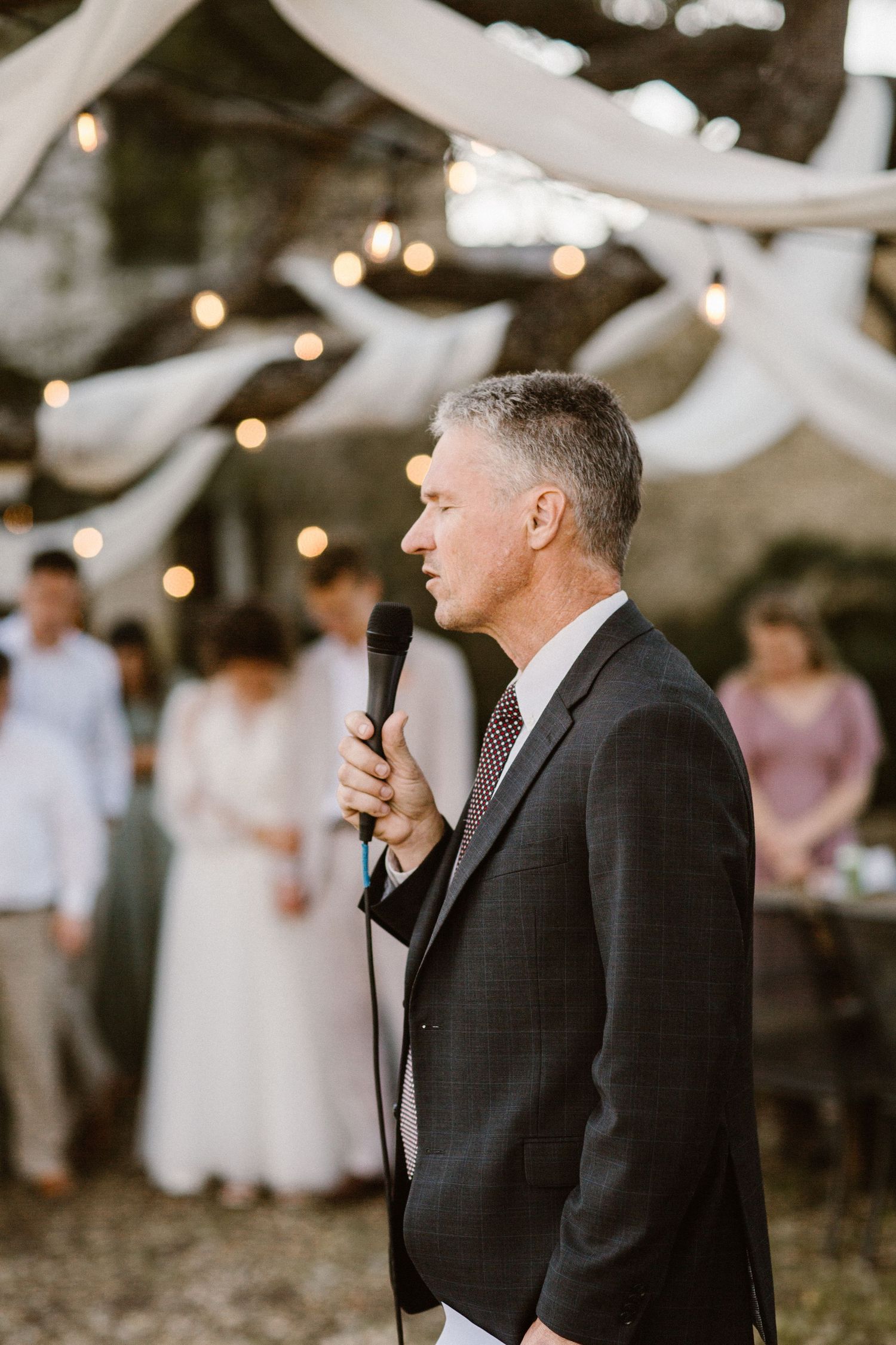 A man in a dark suit delivers a speech with a microphone at an outdoor wedding reception.