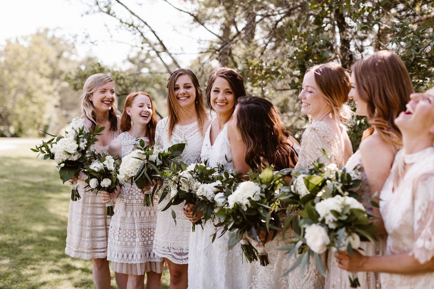 Bride and bridesmaids in white dresses laughing while holding white floral bouquets.
