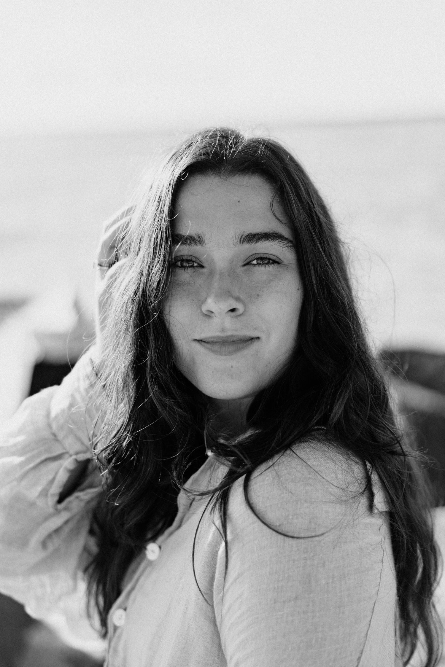 Black and white portrait of a young woman with long hair smiling softly near the ocean.