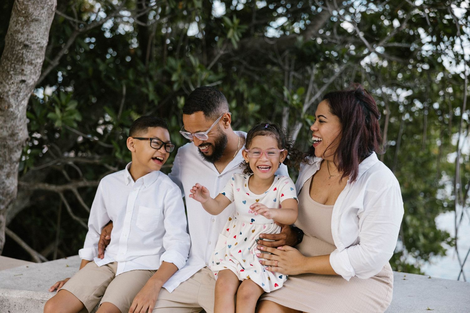 Happy family of four sitting together outdoors near trees, smiling and laughing in coordinated white and beige outfits.