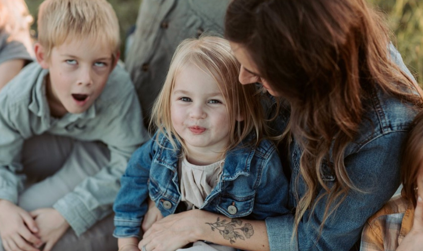 Mother with tattoos smiling at young blonde girl while older boy makes funny face outdoors.