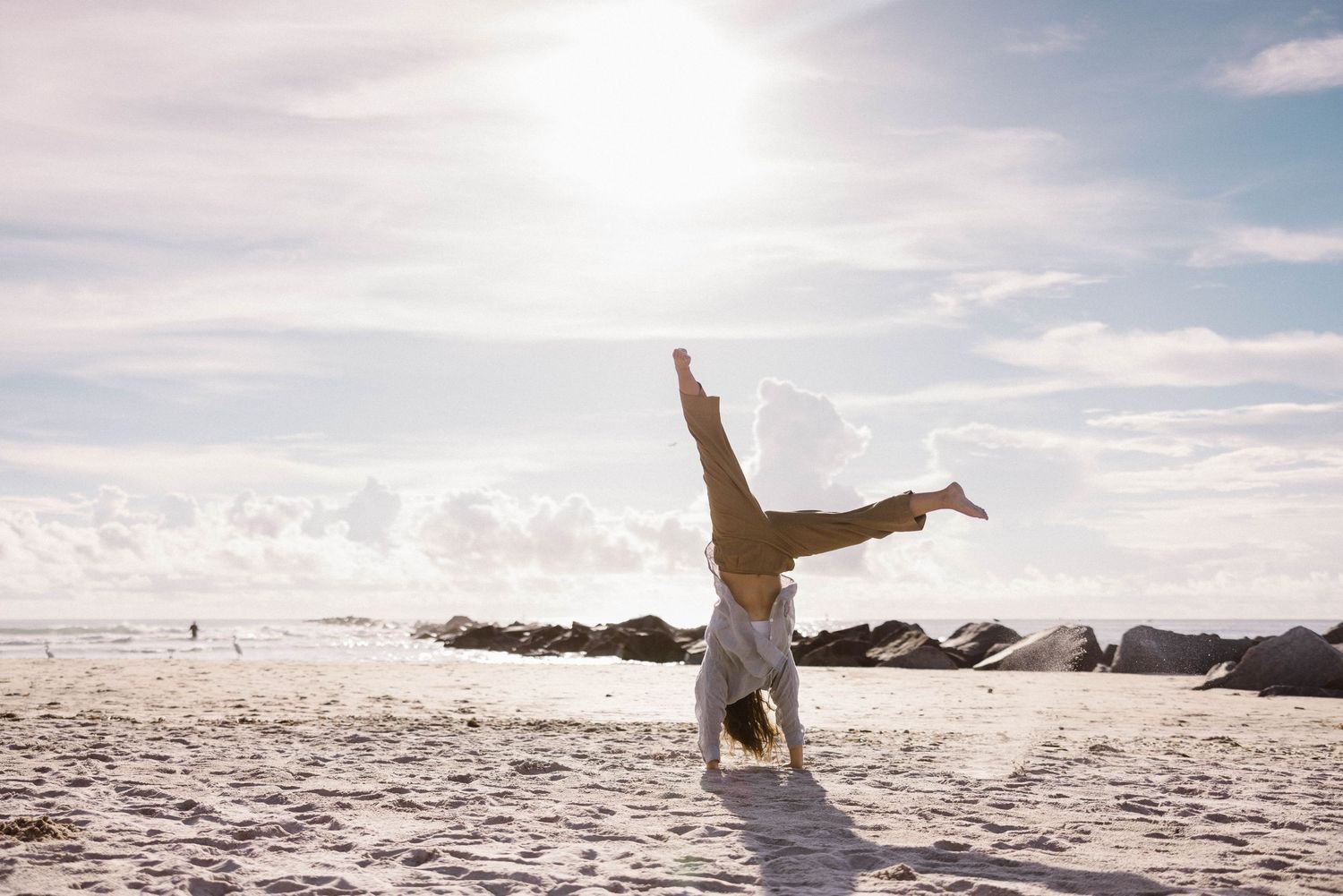 A person does a cartwheel on a sunny white sand beach with rocky shoreline in the background.