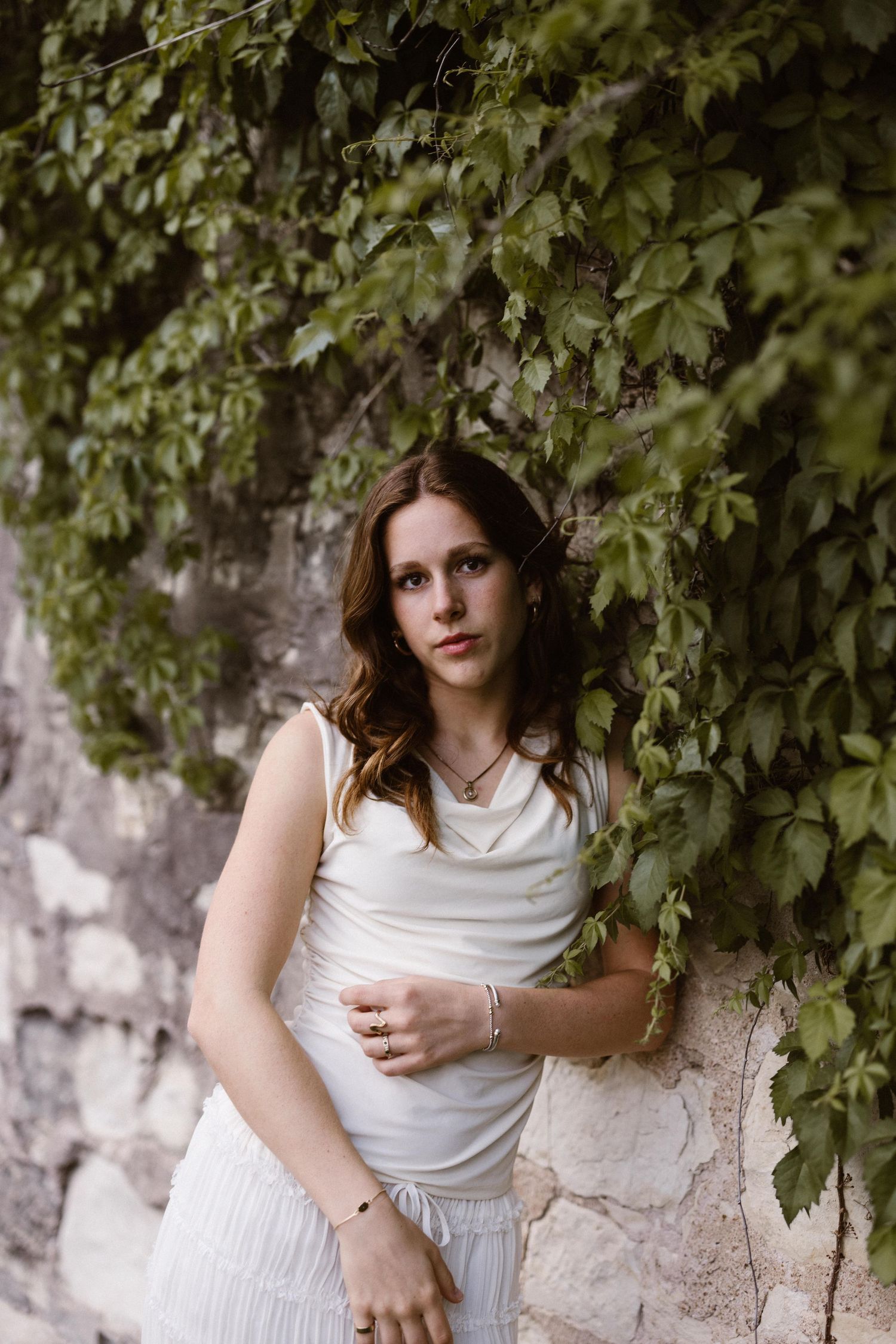 Young woman in a white sleeveless top and skirt standing against a stone wall covered in green ivy.