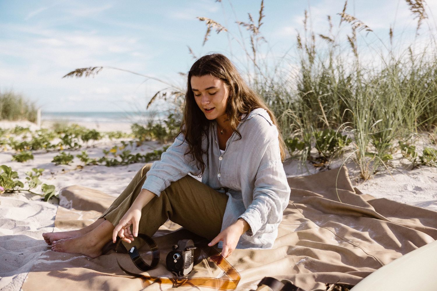 Woman in casual linen outfit sitting on blanket in sandy beach dunes with camera.