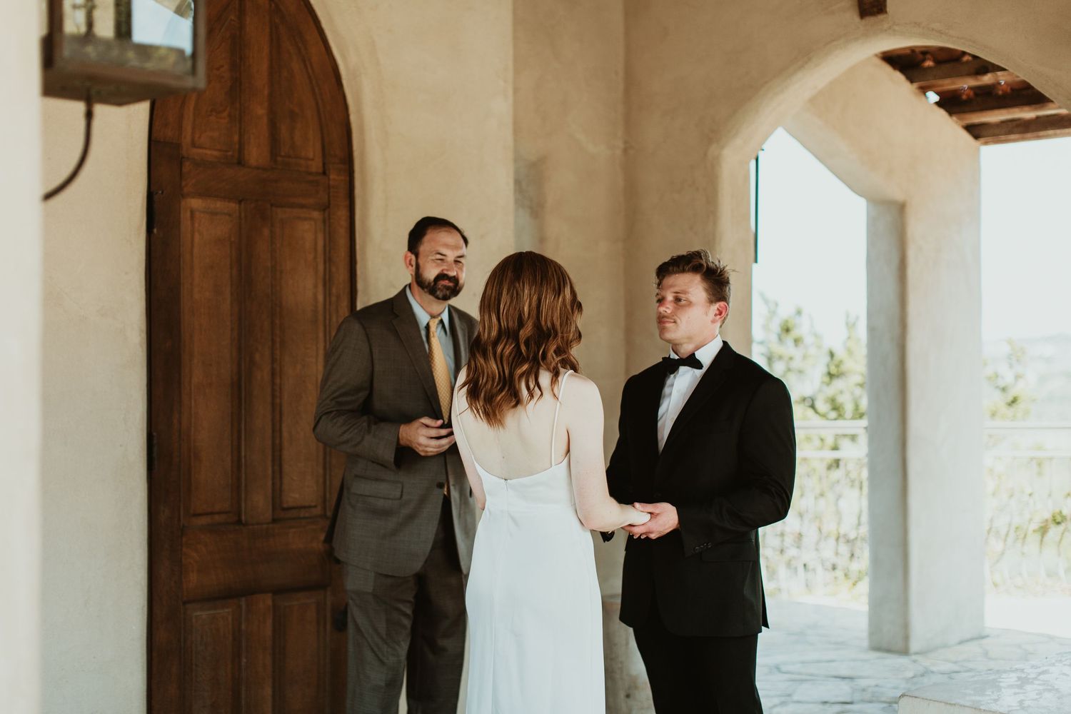Bride and groom exchange vows with officiant at rustic outdoor wedding ceremony near wooden doors.