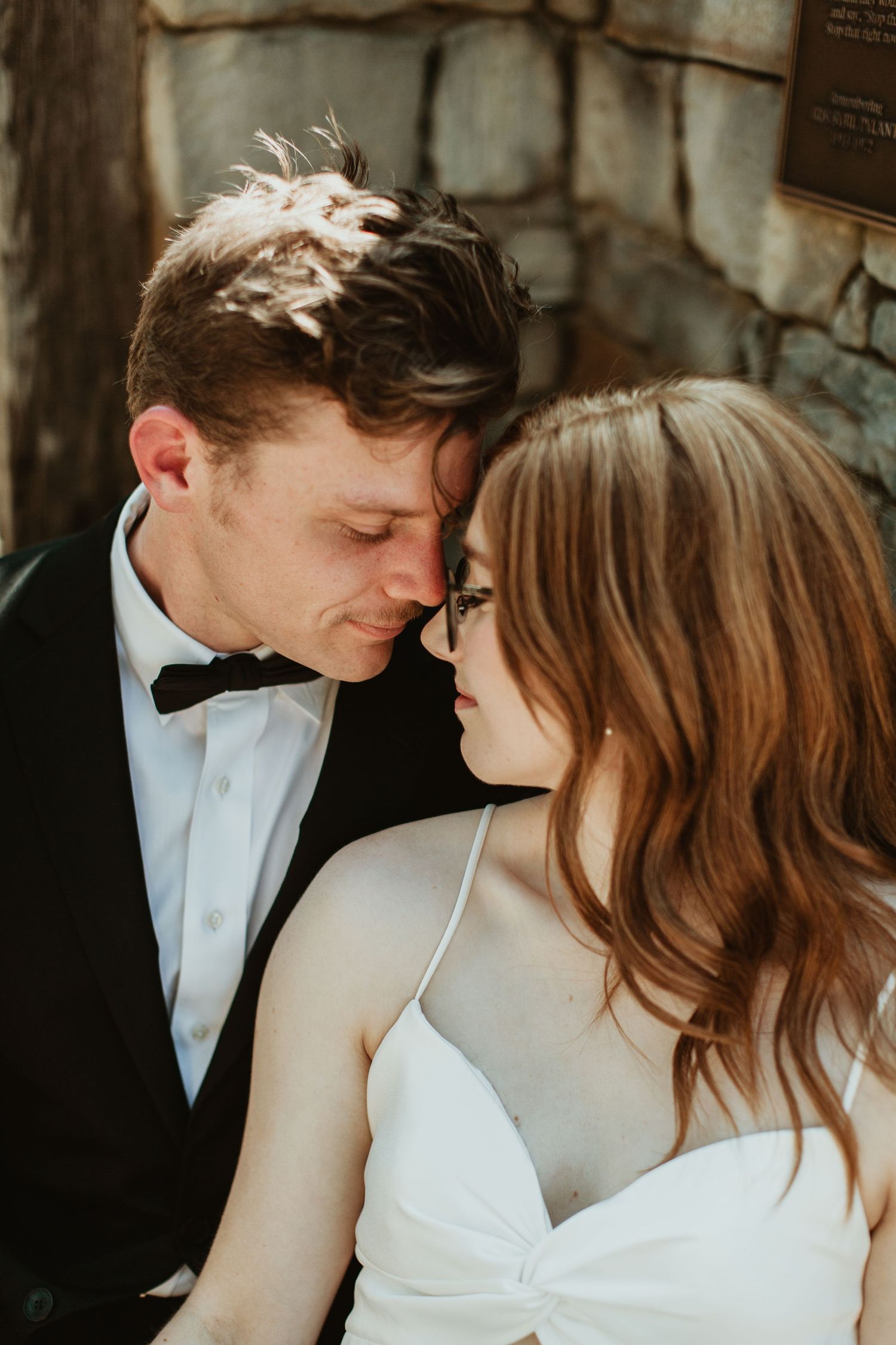 Romantic wedding couple in formal attire share an intimate moment, groom in tuxedo and bride in white dress with glasses.