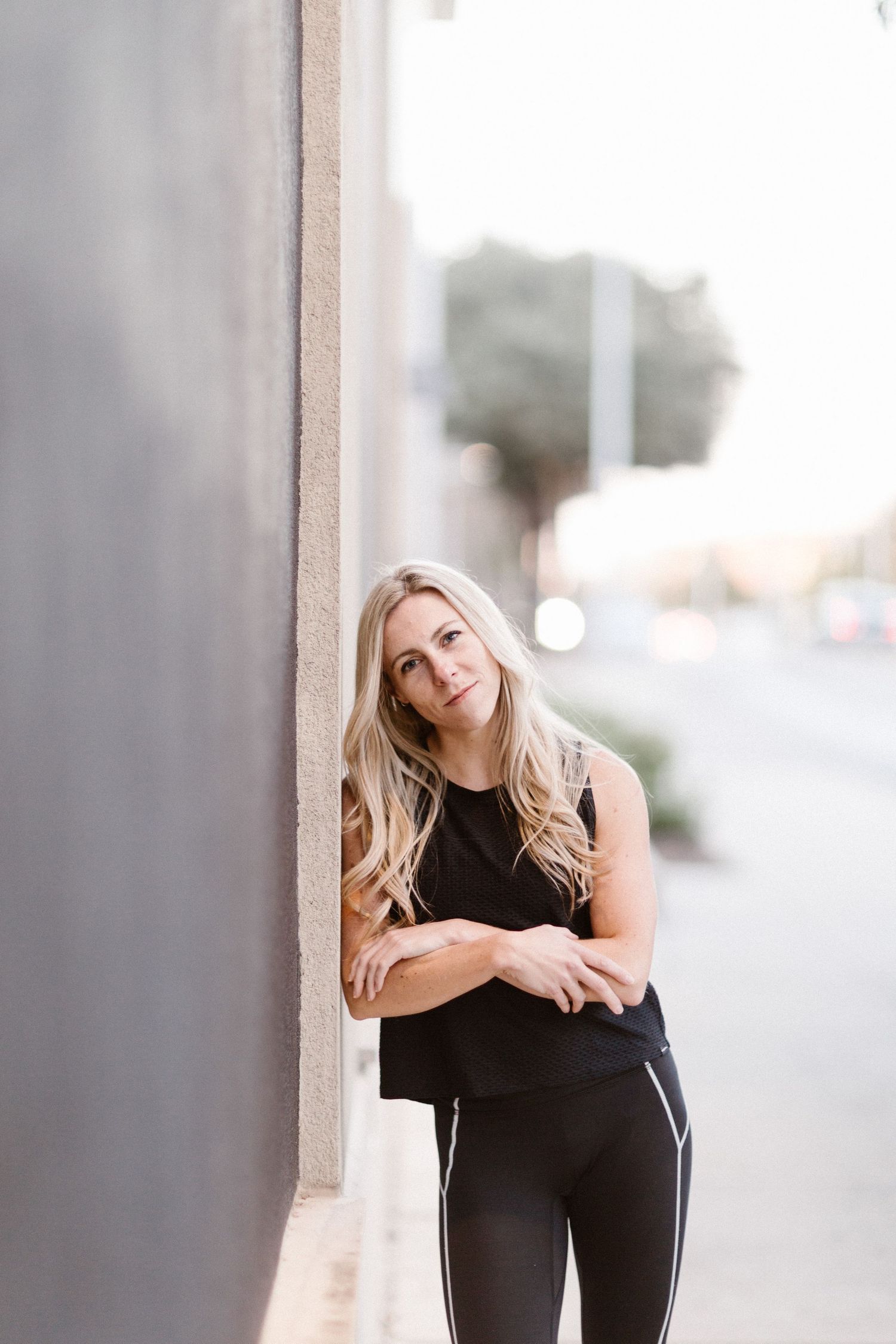 Blonde woman in black athletic wear leaning against a wall outdoors, arms crossed, smiling confidently.