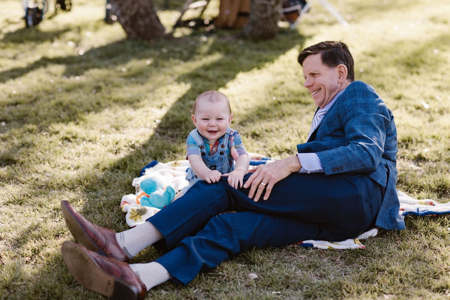 Smiling man in navy suit sits on grass with a happy baby at an outdoor wedding.