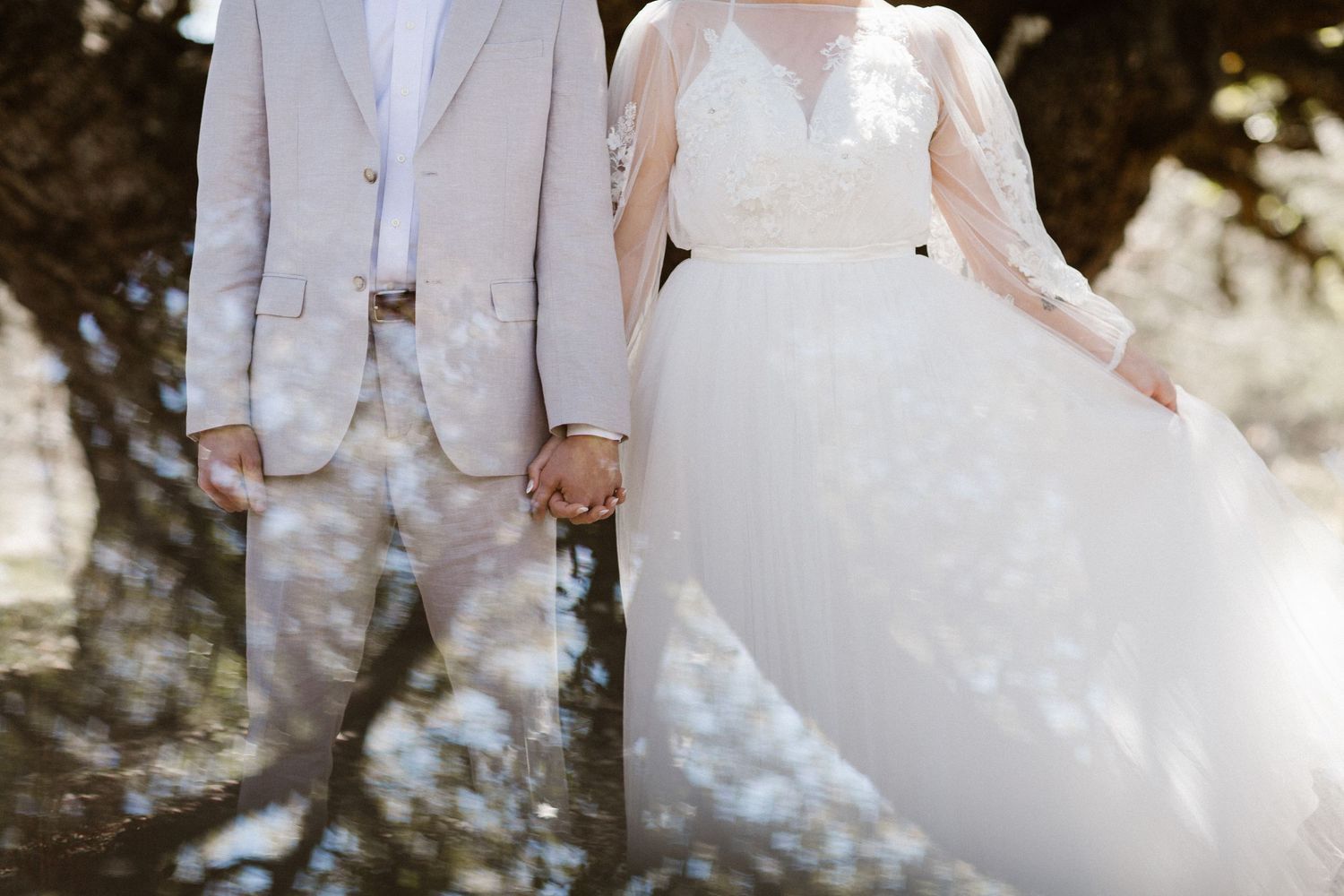 Bride in flowing white lace gown and groom in beige suit hold hands outdoors.