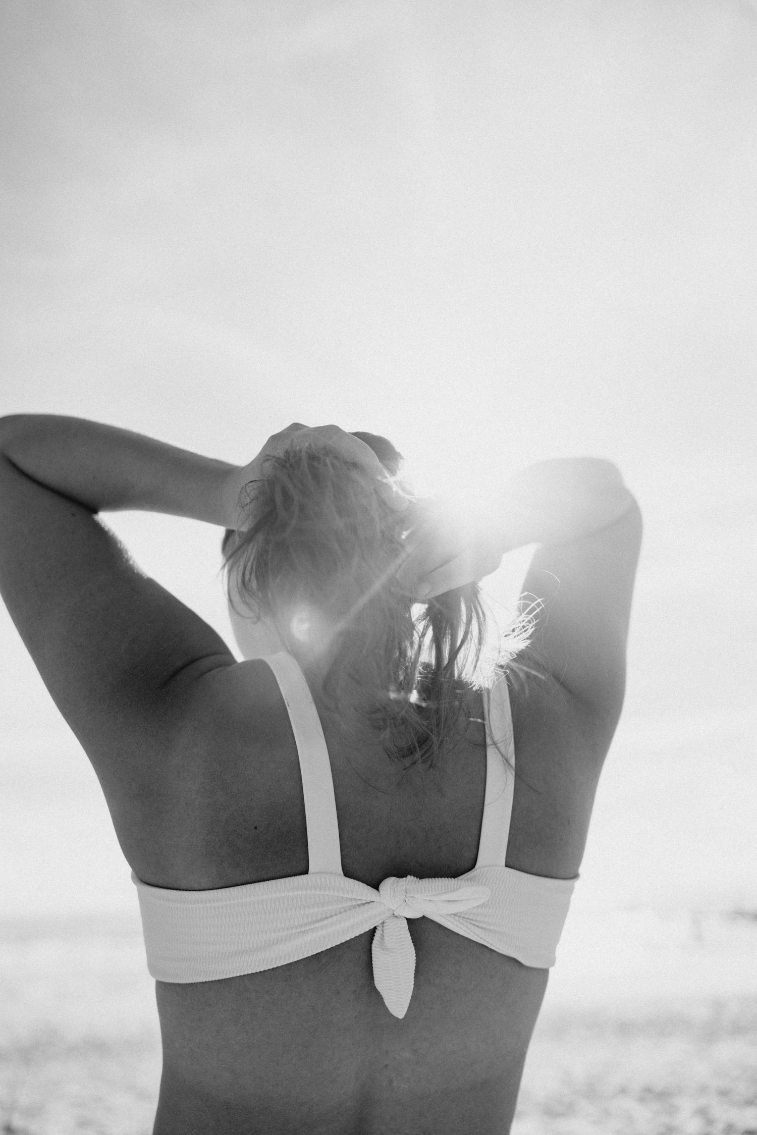 Black and white photo of a woman in a bikini top adjusting her hair with sun flare on a beach.